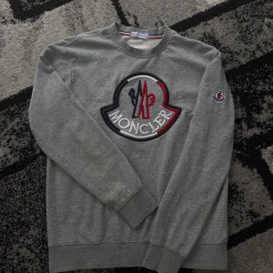 Moncler tröja - Snygg grå sweatshirt från Moncler med stor broderad logga i vitt, rött och blått på bröstet. Tröjan har rund hals, långa ärmar och Moncler-märke på ärmen. Tillverkad i mjuk bomull för en skön och avslappnad stil. Storleken är M men jag skulle sagt S sitter perfekt på mig