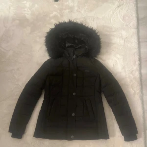 Svart pufferjacka från Marikoo -  pufferjacka från Marikoo med fluffig huva. Jackan har långa ärmar, dragkedja och knappar framtill samt två fickor. Insidan är fodrad perfekt för kalla dagar! Använd en vinter. Där man hänger jackan har jag sytt då den gick sänder bara  nån vecka när jag hade den men har funkat bra att hänga på resten av förra vintern. Såklart kan man sprätta och sy om proffisgare. På fluffet har en knapp gått av men den är näst längst ut så de sitter fof fast men man kan ju sy till en knap. Knappen har jag inte