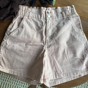 Ljusrosa shorts från KappAhl - Snygga ljusrosa shorts från KappAhl med hög midja och klassisk femficksdesign. De är tillverkade i mjuk bomull med lite stretch för extra komfort. Perfekta för varma dagar och har dubbla knappar framtill samt hällor för bälte.