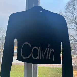 Svart croppad hoodie Calvin Klein - Svart croppad hoodie från Calvin Klein Jeans med stor vit logga över magen. Använd ett fåtal gånger, storlek M-L men jag skulle säga att den är en S eftersom jag vanligtvis har XS/S och den passar mig perfekt .