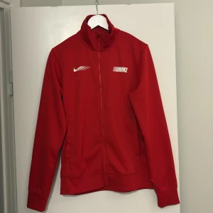 Röd ziptröja från Nike i medium - Snygg röd ziptröja från Nike med vit logga och coolt tryck på bröstet. Tröjan har hög krage, långa ärmar och två sidofickor. Materialet är mjuk polyester som passar perfekt till träning eller chill. Dragkedja hela vägen framtill för enkel på- och avtagning.