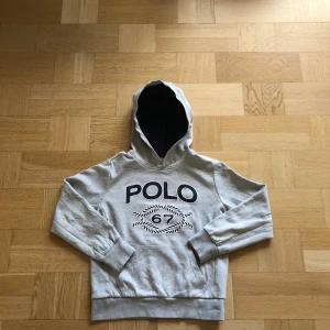 Grå hoodie från Polo Ralph Lauren - Snygg grå hoodie från Polo Ralph Lauren med stor POLO-logga och siffran 67 tryckt på bröstet. Tröjan har huva med mörkblå insida, ribbade muddar och en klassisk känguruficka framtill. Perfekt för chill dagar och streetstyle.