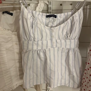 Randig linnetopp från Brandy Melville - Säljer en söt vit linnetopp från Brandy Melville med ljusblå ränder. Toppen har smala axelband, knappar framtill, markerad midja och en snygg knytning i ryggen. Perfekt för varma dagar och har en lätt, luftig känsla tack vare bomullsmaterialet.