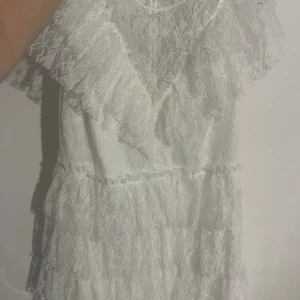 Vit spetsklänning med volanger - Superfin vit klänning i spets med volangdetaljer och pärlknappar i ryggen. Klänningen har kort ärm och flera lager spets som ger en romantisk känsla. Perfekt för sommarens alla tillfällen!