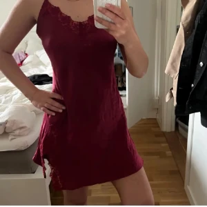 Vinröd spetsklänning Lady Avenue - Vinröd kortklänning från Lady Avenue med tunna axelband och vacker spetsdetalj vid urringningen och längs ena sidan. Klänningen har en slits och är tillverkad i glansigt satinliknande material som känns lyxigt och mjukt mot huden.