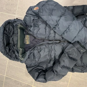 Grå pufferjacka Scotch & Soda - Snygg grå pufferjacka från Scotch & Soda med huva och flera fickor framtill. Jackan har snygg design, dragkedja och coola detaljer . Perfekt för kalla dagar och har en modern stil. En lagning på sidan. se bild 3.