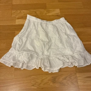 Vit volangkjol från ASOS DESIGN - Supersöt vit kortkjol från ASOS DESIGN med broderade hål och volangkant. Kjolen har en feminin och luftig look, perfekt för varma dagar. Tillverkad i bomull och stängs med dragkedja i sidan. Passformen är normal och tyget är lätt och skönt.