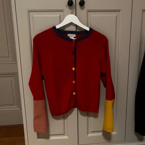 Färgglad kofta från Stella McCartney - Säljer en cool röd kofta från Stella McCartney med kontrastfärgade muddar i rosa och gult. Koftan har blå kant vid halsen och längs knäppningen, samt färgglada knappar. Materialet är mjuk bomull och modellen är långärmad med normal passform.