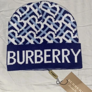 Blå vit Burberry mössa - Snygg mössa från Burberry i blått och vitt med ikoniskt monogrammönster och stor logga framtill. Mössan är stickad och har en bred uppvikt kant. Perfekt accessoar för att lyfta din vinterstil.