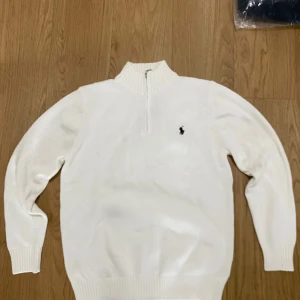Vit stickad tröja Polo Ralph Lauren - Snygg vit stickad tröja från Polo Ralph Lauren med half zip och ribbad krage. Klassisk logga broderad på bröstet i svart. Tröjan har långa ärmar och ribbade muddar vid ärmslut och nederkant. Perfekt för dig som gillar stilrena och tidlösa plagg.