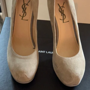 Beige Yves Saint Laurent pumps med klack - Snygga beige pumps från Saint Laurent i mjuk mocka med rund tå och hög klack. Skorna har en diskret plattform och klassisk siluett som ger en elegant look. Perfekta för dig som vill ha lyxiga och stilrena klackar med exklusiv känsla. Klackhöjd 15 cm