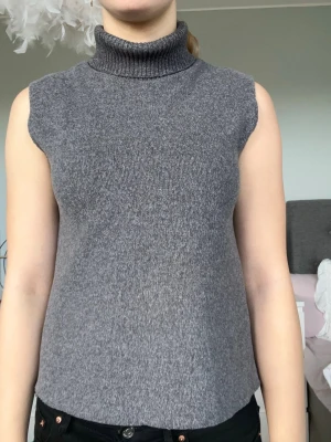 Grå stickad polotopp - Säljer en grå stickad polotopp med hög krage och ärmlös design. Toppen har en mjuk och stretchig känsla, perfekt för lager på lager eller som den är. Enkel och stilren look som passar till det mesta.