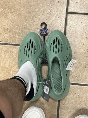 Green yeezy foam runners - Säljer ett par gröna sandaler från mixit i storlek 10. De är tillverkade i lätt gummi med ventilerande hål och slip-on design. Perfekta för chill dagar och enkla att ta på. Modern look med futuristiska detaljer.