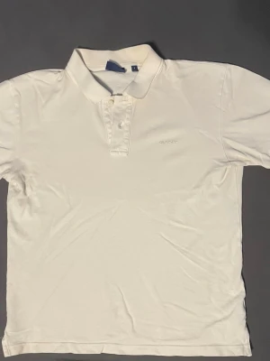 Vit piké från GANT - Klassisk vit pikétröja från GANT med diskret broderad logga på bröstet. Tröjan har krage och knappar framtill för en clean look. Tillverkad i mjuk bomull som känns skön mot huden. Perfekt för dig som gillar stilrena och tidlösa plagg.