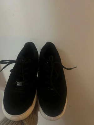 Svarta sneakers med vit sula - Stilrena svarta sneakers med tjock vit sula och svarta skosnören. Ovandelen är i mockaliknande material och har metall-detaljer vid snörningen. Perfekta för en avslappnad streetwear-look.