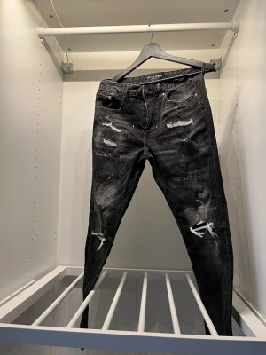Slim fit jeans med slitningar - Svarta jeans med slitna detaljer och flera revor på benen från Supply and  Demand. Modellen har fem fickor, knappgylf och en smal passform som sitter snyggt. Perfekt för dig som gillar en edgy streetstil.