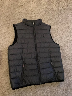  Ralph Lauren väst  - Säljer en svart dunväst från Polo Ralph Lauren i storlek L. Västen har quiltad design, två fickor med dragkedja och hög krage. Perfekt att slänga över en hoodie för en chill look. Klassisk logga på bröstet och stilren passform.