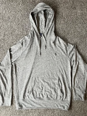 Grå hoodie från Polo Ralph Lauren - Snygg grå hoodie från Polo Ralph Lauren med klassisk huva och dragsko. Tröjan har en broderad logga på bröstet och en stor känguruficka framtill. Tillverkad i mjukt bomullsmaterial och har ribbade muddar vid ärmslut och nederkant.