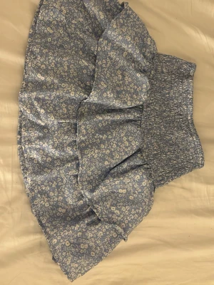 Blommig blå volangkjol Gina Tricot - Supersöt blå kortkjol från Gina Tricot med vitt blommönster och bred smock i midjan. Kjolen har volanger som ger en luftig och lekfull känsla, perfekt för varma dagar. Tillverkad i mjukt och lätt material.