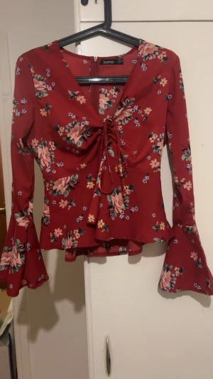 Blommig röd blus med volangärm - Supersöt röd blus från Boohoo med blommigt mönster, v-ringning och snörning framtill. Långa trumpetärmar och volang nertill ger en romantisk vibe. Perfekt att styla med jeans eller kjol för en trendig look. Ganska använd men inget tecken på slitage. Strl 36/S.