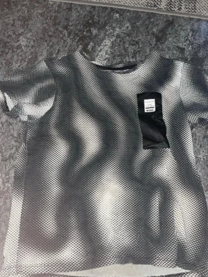 Svartvit mönstrad t-shirt med ficka - Cool svartvit t-shirt från Adidas med unikt vågigt prickmönster och svart bröstficka med logga. T-shirten har rund hals och korta ärmar, perfekt för en trendig streetstyle-look.