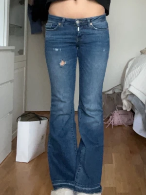 Blå bootcut jeans med slitningar - Säljer ett par blå bootcut jeans med slitningar på framsidan och bakfickor. Lågmidja och utsvängda nertill.