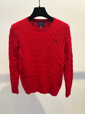 Röd kabelstickad tröja Polo Ralph Lauren - Kabelstickad röd tröja från Polo Ralph Lauren med rund halsringning och broderad logga på bröstet. Tröjan har långa ärmar och ribbade muddar vid ärmslut och nederkant. Perfekt för en snygg och klassisk look.