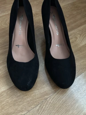 Svarta pumps från Tamaris - Stilrena svarta pumps från Tamaris med rund tå och klädd klack. Skorna har en ovandel i mockaimitation och innersula med Touch it-teknologi för extra komfort. Perfekta för dig som gillar klassisk och tidlös stil. Tyvärr lite avskavt på ena klacken se bild.