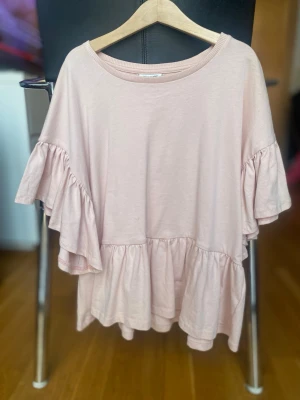 Ljusrosa volangblus med kort ärm - Supersöt ljusrosa blus med volanger på både ärmar och nederkant. Blusen har en lös passform och är tillverkad i mjuk bomull, perfekt för en chill och trendig look. Rund halsringning och korta ärmar med extra volym för en lekfull vibe.Är som xs/s.Använd en gång