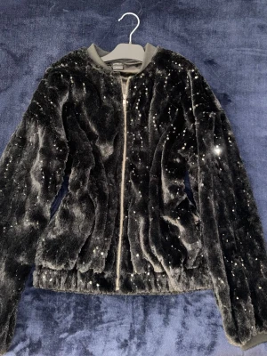 Svart fleecetröja med paljetter - Säljer en svart fleecetröja med glittriga paljettdetaljer. Jackan har dragkedja framtill, rundad krage och är supermjuk i materialet. Perfekt för dig som både vill se snygg ut och hålla sig varm samtidigt.