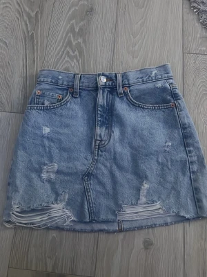 Blå jeanskjol med slitningar - Trendig  fin blå jeanskjol i kort modell med slitna detaljer och rå kant nertill. Kjolen har klassiska fickor fram och bak samt bälteshällor. Använd två gånger men säljs då den är för stor. 