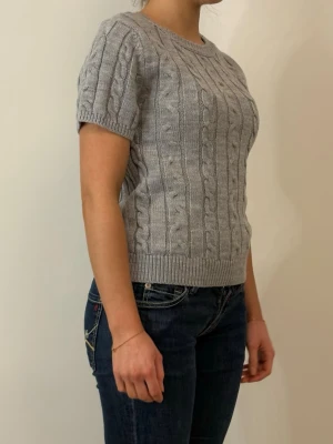 Grå kabelstickad kortärmad tröja - Snygg grå kabelstickad tröja med korta ärmar och rund halsringning. Tröjan har ribbstickade muddar vid ärmslut och nederkant. Perfekt för lager-på-lager och passar till både jeans och kjol.