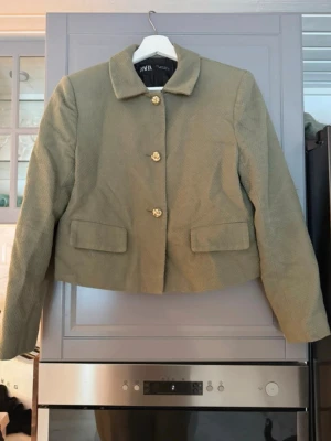 Olivgrön kavaj från Zara - Stilren olivgrön kavaj från Zara med klassisk krage och tre dekorativa guldfärgade knappar framtill. Kavajen har två fickor med lock och är tillverkad i ett strukturerat tyg som ger en exklusiv känsla. Perfekt för dig som gillar en trendig och elegant look.