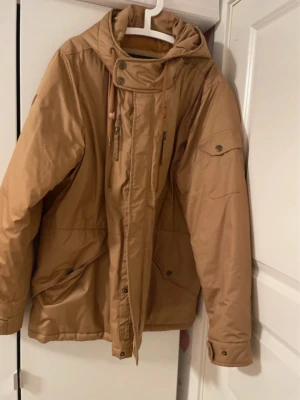 Beige herr parkasjacka med huva, 2XL - Snygg beige parkasjacka med dragkedja, knappar och justerbar huva. Jackan har flera fickor med dragkedja och knappstängning samt hög krage med snörning. Perfekt för kyliga dagar och enkel att matcha med olika outfits.