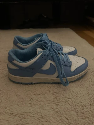 Nike Dunk Low blå och vita sneakers - Snygga Nike Dunk Low sneakers i blått och vitt. Skorna har klassisk låg siluett, perforerad tå och matchande blå skosnören. Ovandelen är i läder med vit bas och ljusblå detaljer, samt ikoniska swoosh-loggan på sidorna. Perfekta för en streetwear-look.