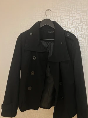 Svart kapp från Vila med knappar - Stilren svart kappa från Vila med dubbelknäppning och knappar framtill. Kappan har bred krage, axelklaffar och är helfodrad för extra komfort. Perfekt för dig som vill ha en klassisk och snygg look under kallare dagar. Den är köpt på stockhomsstadsmission så den är såklart använd men den har inga defekter alls den är i storlek M men passar även S