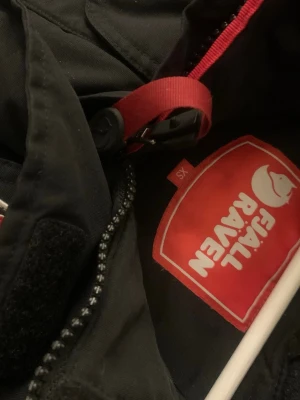 Svart parkas från Fjällräven XS - Svart parkasjacka från Fjällräven med dragkedja, stora fickor framtill och klassisk röd logotyp på bröstet. Jackan har huva med röd detalj och är perfekt för kyliga dagar. Snygg och funktionell design.