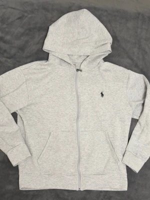 Ljusgrå hoodie från Polo Ralph Lauren - Ljusgrå hoodie med dragkedja från Polo Ralph Lauren. Klassisk design med huva, två fickor framtill och den ikoniska lilla svarta logotypen broderad på bröstet. Tillverkad i mjukt bomullsmaterial som känns skönt mot huden. Perfekt för en avslappnad och stilren look.