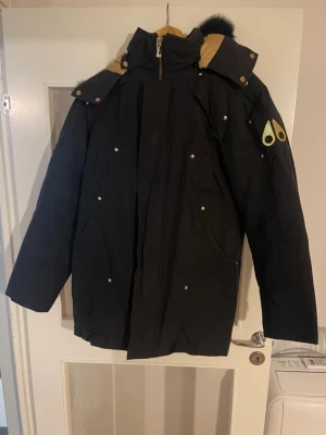 Svart parkas från Moose Knuckles - Svart parkas från Moose Knuckles med guldiga detaljer och logga på ärmen. Jackan har huva med svart fuskpäls, dragkedja och tryckknappar framtill samt flera fickor. Perfekt för kalla dagar med sin långa modell och rejäla material.
