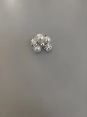 Silverfärgad ring med pärlor och stenar - Unik silverfärgad ring dekorerad med vita pärlor och gnistrande stenar i klusterdesign. Ringen har en öppen justerbar ringbas och en modern, iögonfallande look som verkligen sticker ut.
