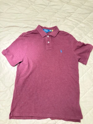 Vinröd pikétröja från Polo Ralph Lauren - Klassisk vinröd pikétröja från Polo Ralph Lauren med blå broderad logga på bröstet. Tröjan har korta ärmar, krage och knappar framtill. Perfekt för en avslappnad men ändå stilren look.