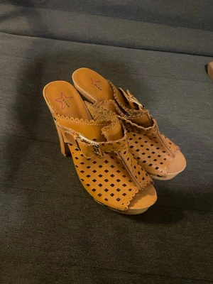 Bruna klackpumps med utskurna detaljer - Snygga bruna pumps från Star med hög klack och utskurna mönster i lädret. Skorna har dekorativa spännen och nitar samt en träliknande sula. Perfekta för dig som gillar unika detaljer och en bohemisk stil.