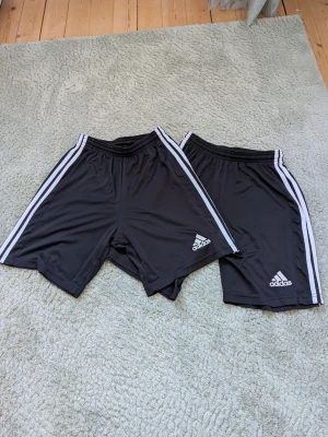Svarta Adidas shorts med vita ränder - Två par svarta Adidas shorts med klassiska vita ränder längs sidorna och Adidas-logga framtill. Tillverkade i lätt och bekvämt polyestermaterial, perfekta för träning eller fotboll. Elastisk midja för skön passform.
