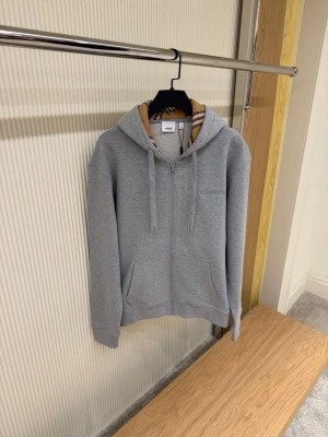 Grå zip hoodie från Burberry - Snygg grå hoodie med dragkedja från Burberry. Klassisk huva med det ikoniska Burberry-rutiga fodret och dragsnören. Två fickor framtill och diskret Burberry-logga broderad på bröstet. Perfekt för en avslappnad och trendig look.