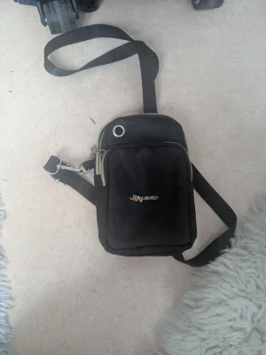 Svart axelväska  - Svart axelväska med justerbar axelrem och två dragkedjefack. Väskan har en metallögla på framsidan och ett litet metallogo(som är lite sönder). Tillverkad i slitstarkt tygmaterial, perfekt för att bära det viktigaste.