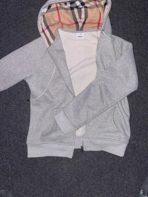 Grå zip-hoodie från Burberry - Snygg grå hoodie från Burberry med dragkedja och klassiskt rutigt foder i huvan. Broderad logga på bröstet och två fickor framtill. Mjuk bomullsblandning som är skön att ha på sig. Perfekt för en avslappnad och stilren look.