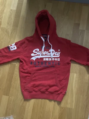 Röd Superdry hoodie med tryck - Röd hoodie från Superdry med vit och blå logga och japanska tecken på bröstet. Stor ficka framtill, vit dragsko i huvan och tryckt siffra på ärmen. Mjuk insida och klassisk passform, perfekt för en avslappnad stil.