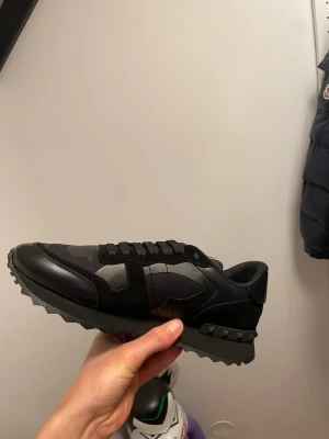 Svarta sneakers från Valentino - Snygga svarta sneakers från Valentino med coola nitar på hälen och camo-detaljer i materialmix av mocka och textil. Skorna har en chunky sula och klassisk låg profil med svarta skosnören. Perfekta för dig som gillar streetstyle och exklusiv design.