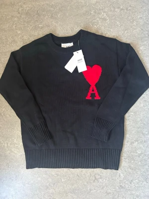 Svart stickad tröja från Ami Paris - Svart stickad tröja från Ami Paris med röd hjärtlogga och bokstaven A på bröstet. Tröjan har rund halsringning, ribbade muddar och nederkant. Perfekt för dig som gillar stilrena plagg med en twist.