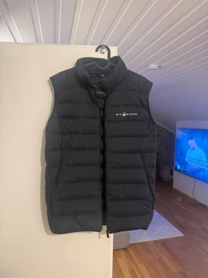 Svart dunväst från Sail Racing - Snygg svart dunväst från Sail Racing med diskret logga på bröstet. Västen har en klassisk quiltad design och hög krage som skyddar mot vind. Perfekt att slänga över en hoodie eller tröja för extra värme under kyliga dagar. Använd max 2 gånger så den är som ny. Passar både tjejer och killar. Storlek 176 men passar mig som har S-M. 